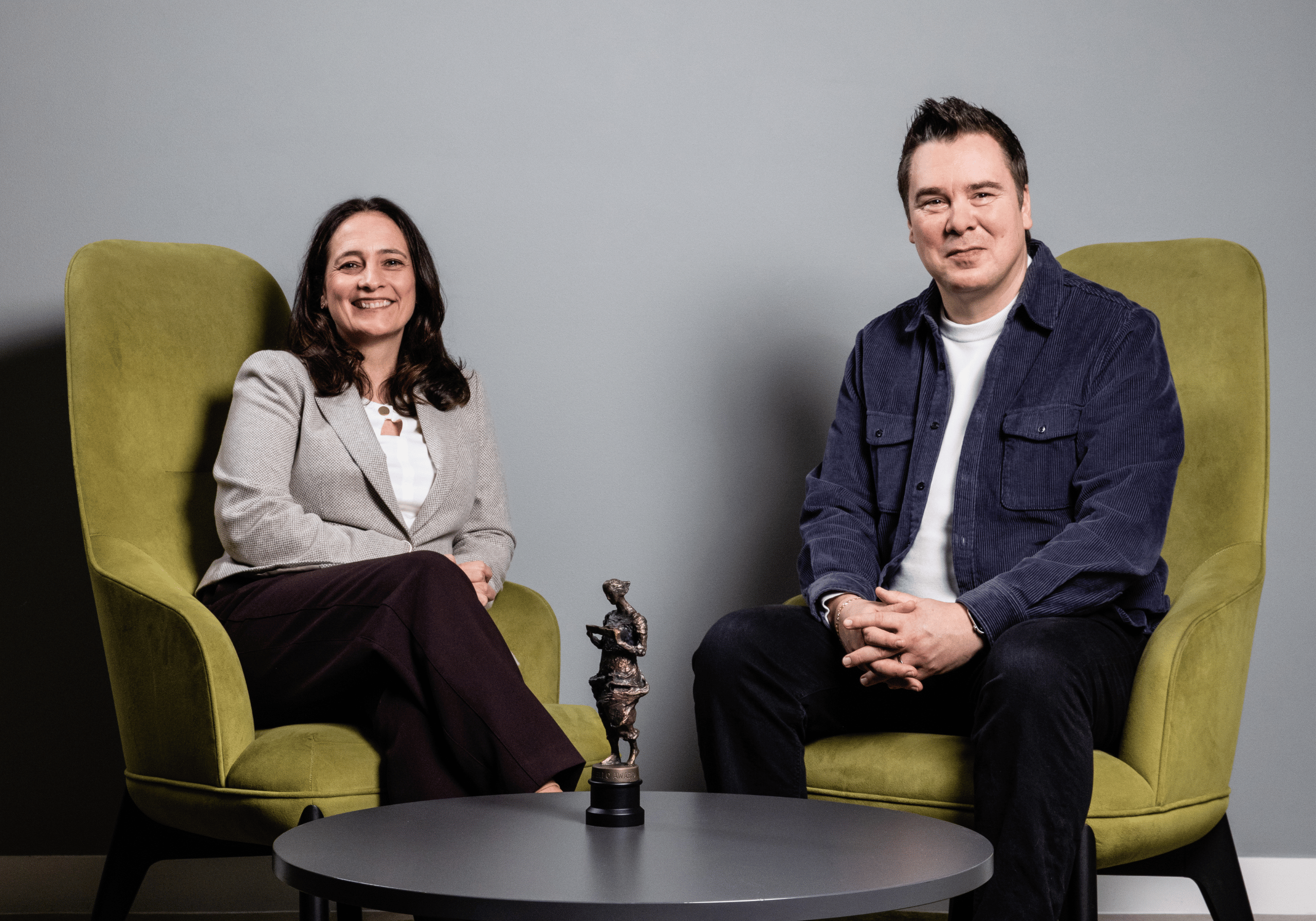 Catherine Martin and Roberto Neri sat on green chairs with Ivor Novello Award on table in front of them