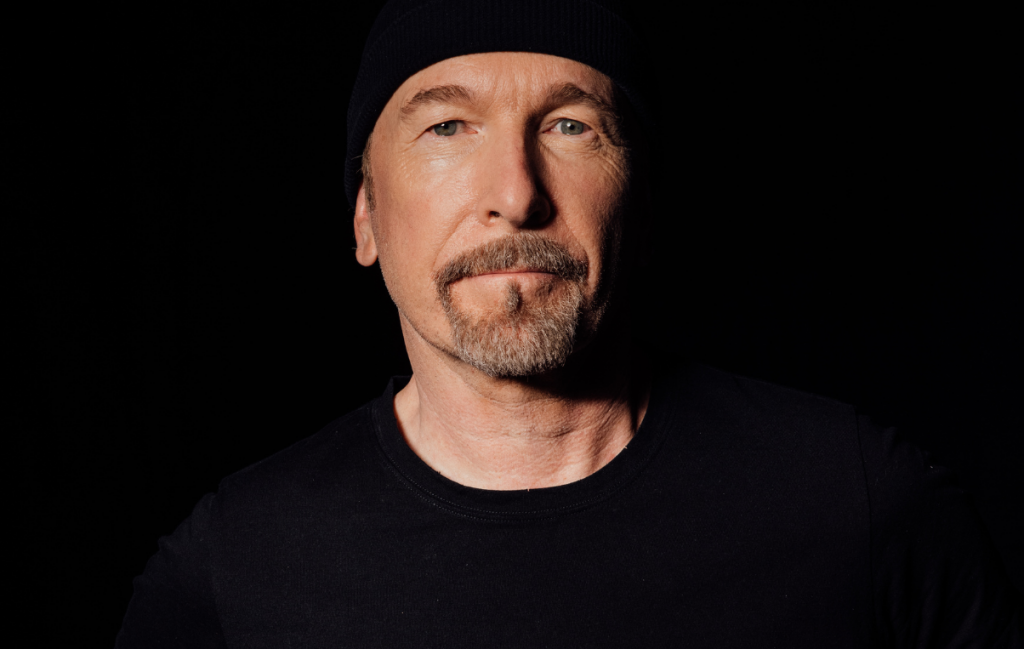 U2's The Edge wearing black t-shirt and hat in front of a dark background
